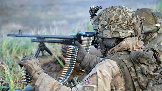 Soldier in desert camouflage gear and helmet feeding a belt-fed machine gun in tall grass.