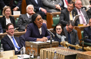 Badenoch offers support to fund warfare through welfare cuts Politician in a purple blazer speaks into a microphone at a ceremonial desk in a parliamentary chamber, with lawmakers watching.