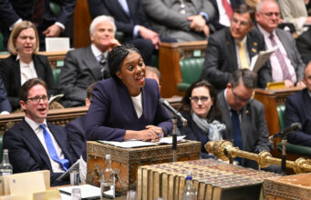 Badenoch offers support to fund warfare through welfare cuts Politician in a purple blazer speaks into a microphone at a ceremonial desk in a parliamentary chamber, with lawmakers watching.