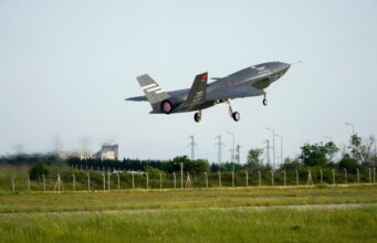 Italian jets fly with Turkish drones in K-SWARM live trial Stealth fighter jet lifting off from a runway, low above a grassy field with a fence in the foreground.