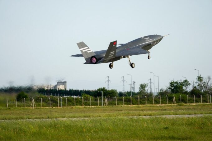 Stealth fighter jet lifting off from a runway, low above a grassy field with a fence in the foreground.