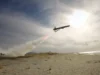 Air tanker plane releasing red retardant over a sandy beach as smoke rises from a ground fire nearby