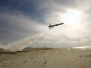 Air tanker plane releasing red retardant over a sandy beach as smoke rises from a ground fire nearby