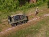 How IDV builds robots that can be reborn as tech advances Drone view of a military convoy-style vehicle on a dirt path with a soldier in camouflage standing nearby among grass and trees.