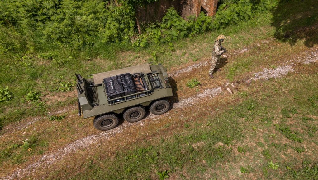 IDV - Viking 2.4 Drone view of a military convoy-style vehicle on a dirt path with a soldier in camouflage standing nearby among grass and trees.