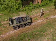 How IDV builds robots that can be reborn as tech advances Drone view of a military convoy-style vehicle on a dirt path with a soldier in camouflage standing nearby among grass and trees.