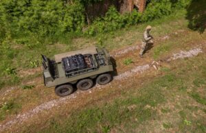 How IDV builds robots that can be reborn as tech advances Drone view of a military convoy-style vehicle on a dirt path with a soldier in camouflage standing nearby among grass and trees.