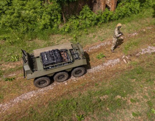 Drone view of a military convoy-style vehicle on a dirt path with a soldier in camouflage standing nearby among grass and trees.
