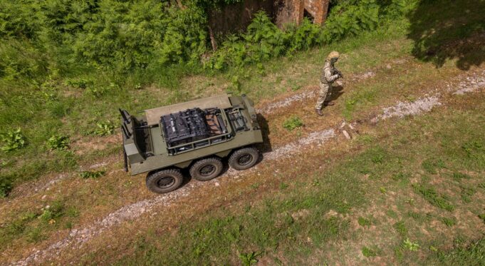 How IDV builds robots that can be reborn as tech advances Drone view of a military convoy-style vehicle on a dirt path with a soldier in camouflage standing nearby among grass and trees.
