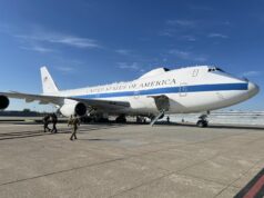 Air Force One parked on a sunny tarmac with stairs deployed to the door and crew nearby
