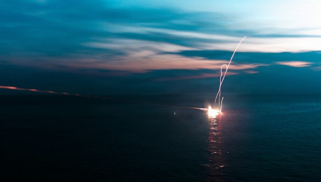 11th MEU Marines, Sailors Conduct Defense of the Amphibious Task Force Live-Fire Exercise Aboard USS Portland Firework explosion on the water at dusk, long-exposure light trail over the dark sea.
