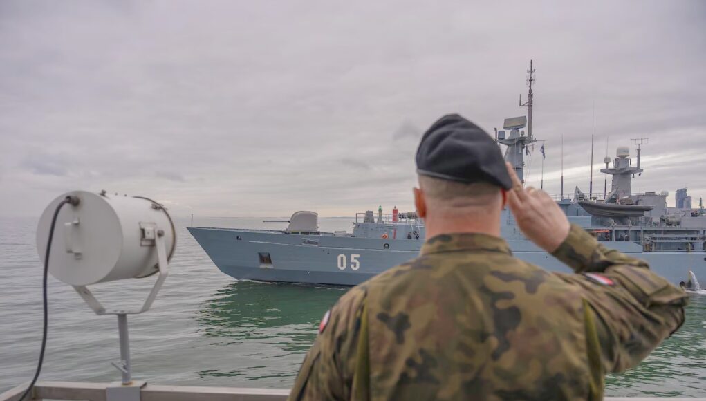 BALTOPS Sailor in camouflage uniform saluting as a gray warship with the number 05 passes by on the water.