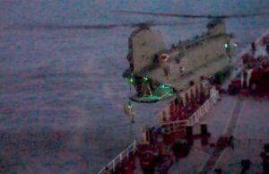 Blurred night photo of a seafront pier with a concrete extension over water and green lights along its edge.