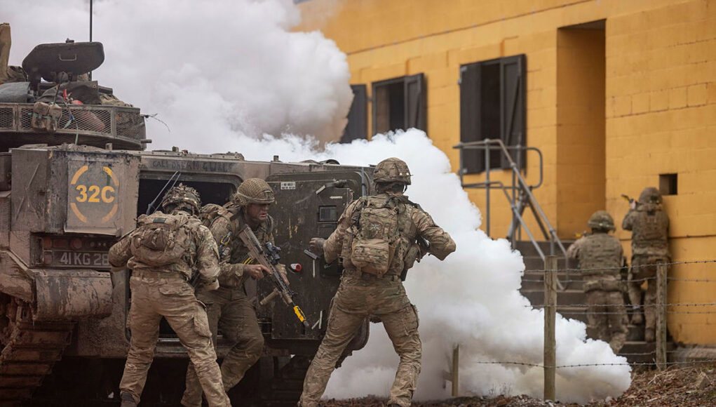 Soldiers in camouflage deploy from an armored vehicle as white smoke billows nearby.