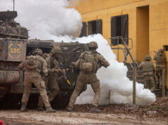 Soldiers in camouflage deploy from an armored vehicle as white smoke billows nearby.