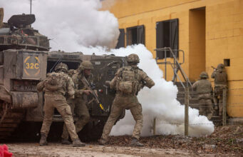 Soldiers in camouflage deploy from an armored vehicle as white smoke billows nearby.