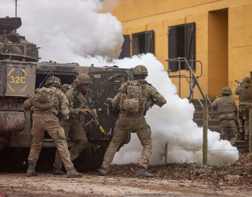 Soldiers in camouflage deploy from an armored vehicle as white smoke billows nearby.