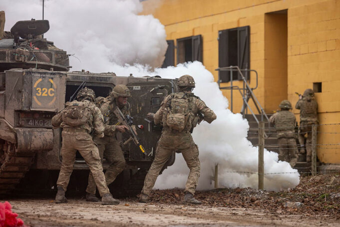Soldiers in camouflage deploy from an armored vehicle as white smoke billows nearby.