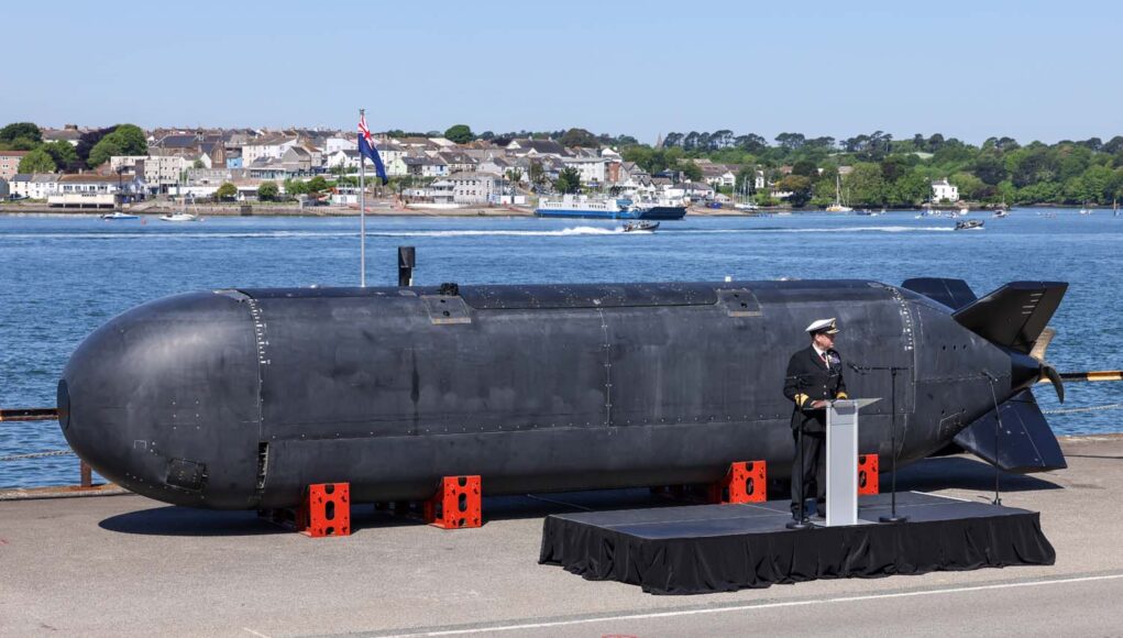 Large black submarine hull displayed on a pier, with a naval officer giving a speech at a podium nearby; harbor and town in the background.