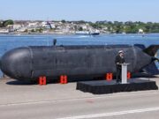 Large black submarine hull displayed on a pier, with a naval officer giving a speech at a podium nearby; harbor and town in the background.