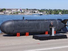 Large black submarine hull displayed on a pier, with a naval officer giving a speech at a podium nearby; harbor and town in the background.