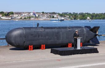 Large black submarine hull displayed on a pier, with a naval officer giving a speech at a podium nearby; harbor and town in the background.