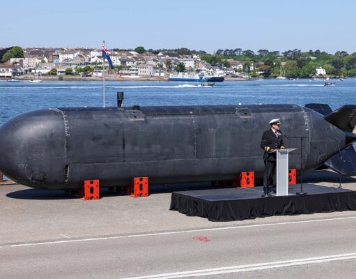 Large black submarine hull displayed on a pier, with a naval officer giving a speech at a podium nearby; harbor and town in the background.