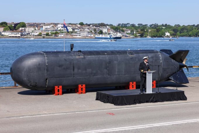 Large black submarine hull displayed on a pier, with a naval officer giving a speech at a podium nearby; harbor and town in the background.