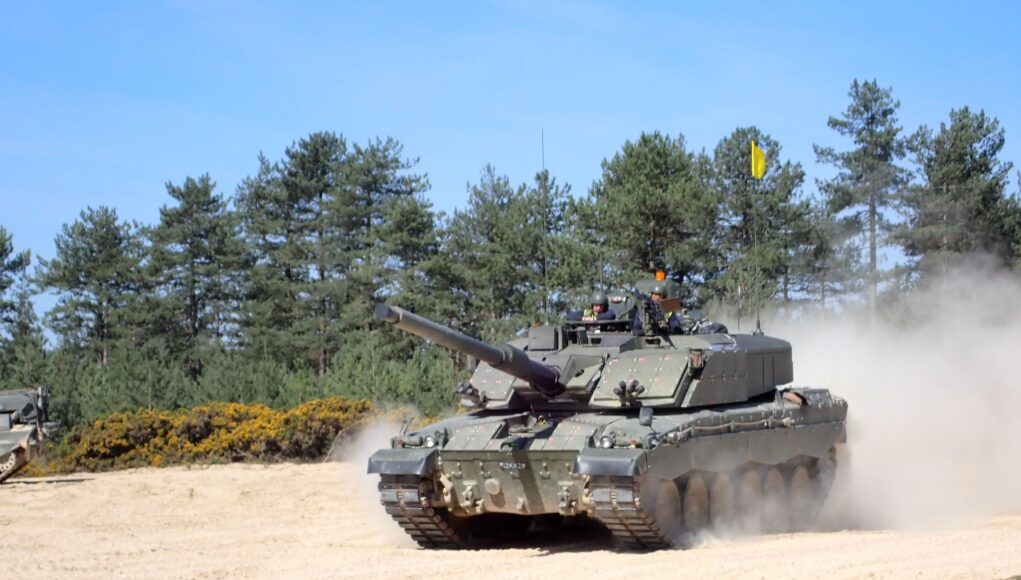 CHALLENGER3rbsl Tank advancing on a sandy field with dust, crew visible in the turret, evergreen trees in the background.