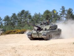 Tank advancing on a sandy field with dust, crew visible in the turret, evergreen trees in the background.