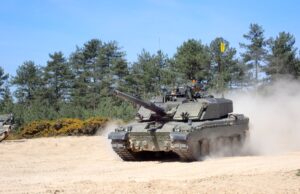 Britain’s new battle tank pushes through trials Tank advancing on a sandy field with dust, crew visible in the turret, evergreen trees in the background.