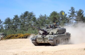 Britain’s new battle tank pushes through trials Tank advancing on a sandy field with dust, crew visible in the turret, evergreen trees in the background.