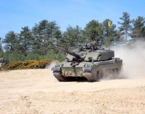 Tank advancing on a sandy field with dust, crew visible in the turret, evergreen trees in the background.