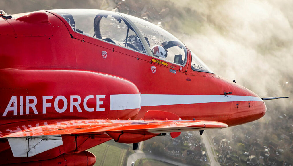 Red aerobatic aircraft in flight with a white side stripe and cockpit canopy, over a rural town and fields below.