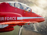 Red aerobatic aircraft in flight with a white side stripe and cockpit canopy, over a rural town and fields below.