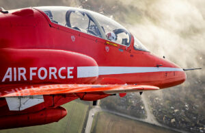 Red Arrows Hawk T1 fleet running low on fatigue life Red aerobatic aircraft in flight with a white side stripe and cockpit canopy, over a rural town and fields below.