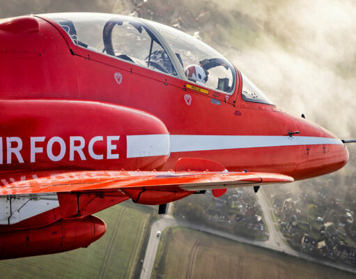 Red aerobatic aircraft in flight with a white side stripe and cockpit canopy, over a rural town and fields below.