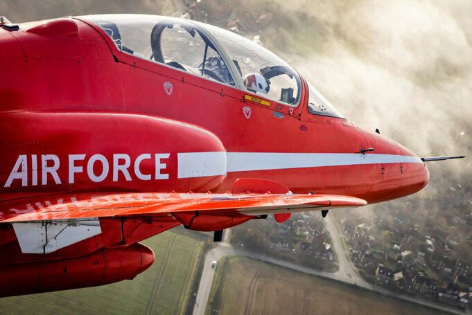 Red Arrows Hawk T1 fleet running low on fatigue life Red aerobatic aircraft in flight with a white side stripe and cockpit canopy, over a rural town and fields below.