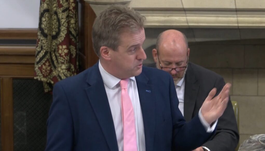 DOWNIEmain Man in a navy suit and pink tie speaks, gesturing with his right hand in a formal meeting; a second man in a suit with glasses sits behind him looking downward.
