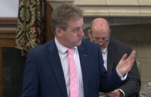 Trust the public on threat resilience, Downie urges Man in a navy suit and pink tie speaks, gesturing with his right hand in a formal meeting; a second man in a suit with glasses sits behind him looking downward.