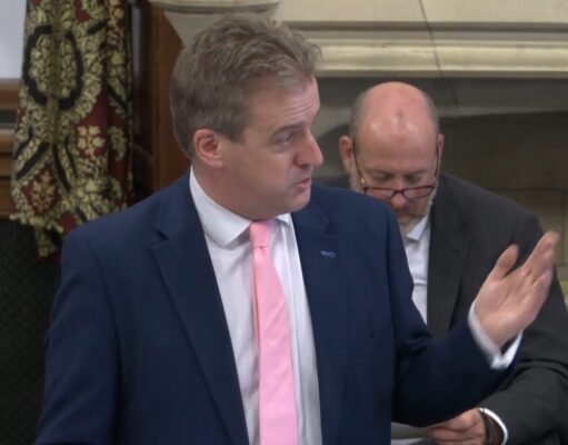 Trust the public on threat resilience, Downie urges Man in a navy suit and pink tie speaks, gesturing with his right hand in a formal meeting; a second man in a suit with glasses sits behind him looking downward.