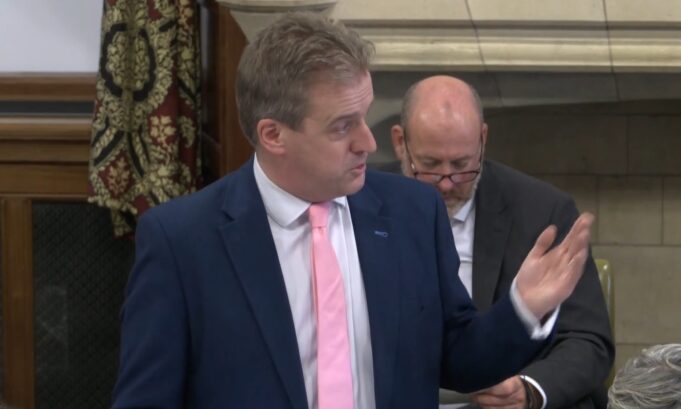 Man in a navy suit and pink tie speaks, gesturing with his right hand in a formal meeting; a second man in a suit with glasses sits behind him looking downward.