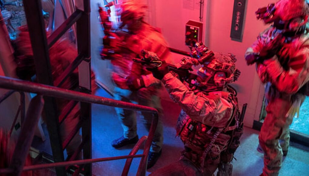 Special-ops style operator in full gear pointing a weapon through a doorway in a dim, red-and-blue lit stairwell.