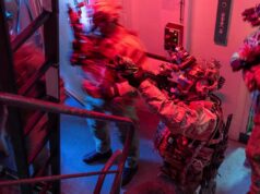 British forces board Russian shadow fleet tanker near UK Special-ops style operator in full gear pointing a weapon through a doorway in a dim, red-and-blue lit stairwell.
