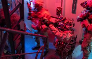 British forces board Russian shadow fleet tanker near UK Special-ops style operator in full gear pointing a weapon through a doorway in a dim, red-and-blue lit stairwell.