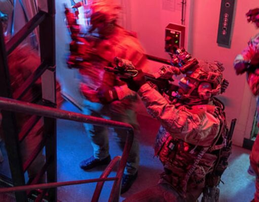 Special-ops style operator in full gear pointing a weapon through a doorway in a dim, red-and-blue lit stairwell.