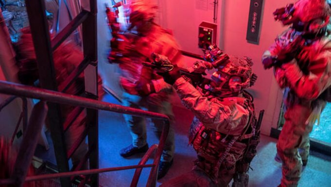 British forces board Russian shadow fleet tanker near UK Special-ops style operator in full gear pointing a weapon through a doorway in a dim, red-and-blue lit stairwell.