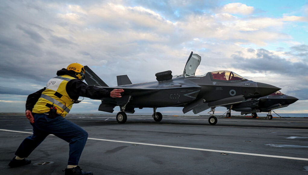 FLEET-20260612-XN0416-176.jpg.iCe_g5uCMjln8wNlYBMA.wohPTJTIQD Crew member in a yellow safety vest pushes a grey F-35 fighter on a crowded aircraft carrier deck under a cloudy sky, cockpit canopy open nearby.