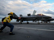 Crew member in a yellow safety vest pushes a grey F-35 fighter on a crowded aircraft carrier deck under a cloudy sky, cockpit canopy open nearby.
