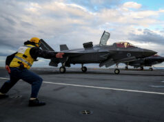 British aircraft carrier launches fighter jets in Arctic Crew member in a yellow safety vest pushes a grey F-35 fighter on a crowded aircraft carrier deck under a cloudy sky, cockpit canopy open nearby.